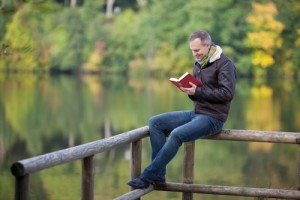 Man Reading Book While Sitting On Fence Against Lake